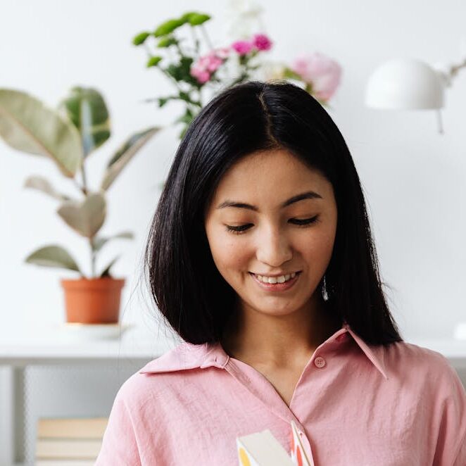 A smiling young Asian woman in a pink shirt reads a colorful book in a home office with plants.