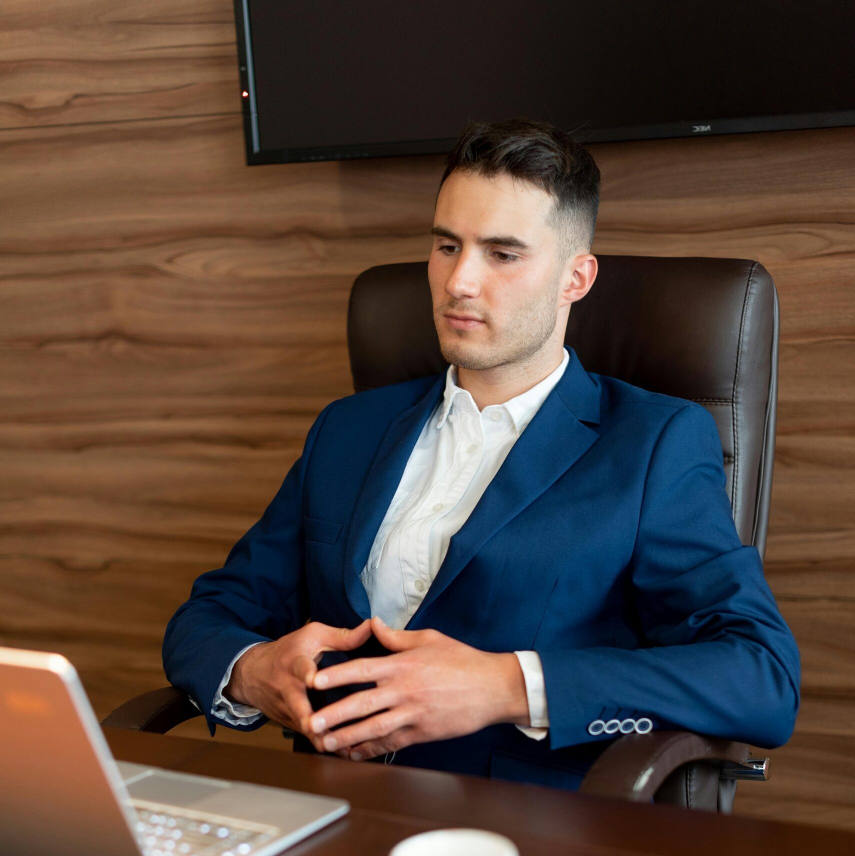 Professional businessman in a blue suit working at a desk with a laptop and coffee in a modern office setting.
